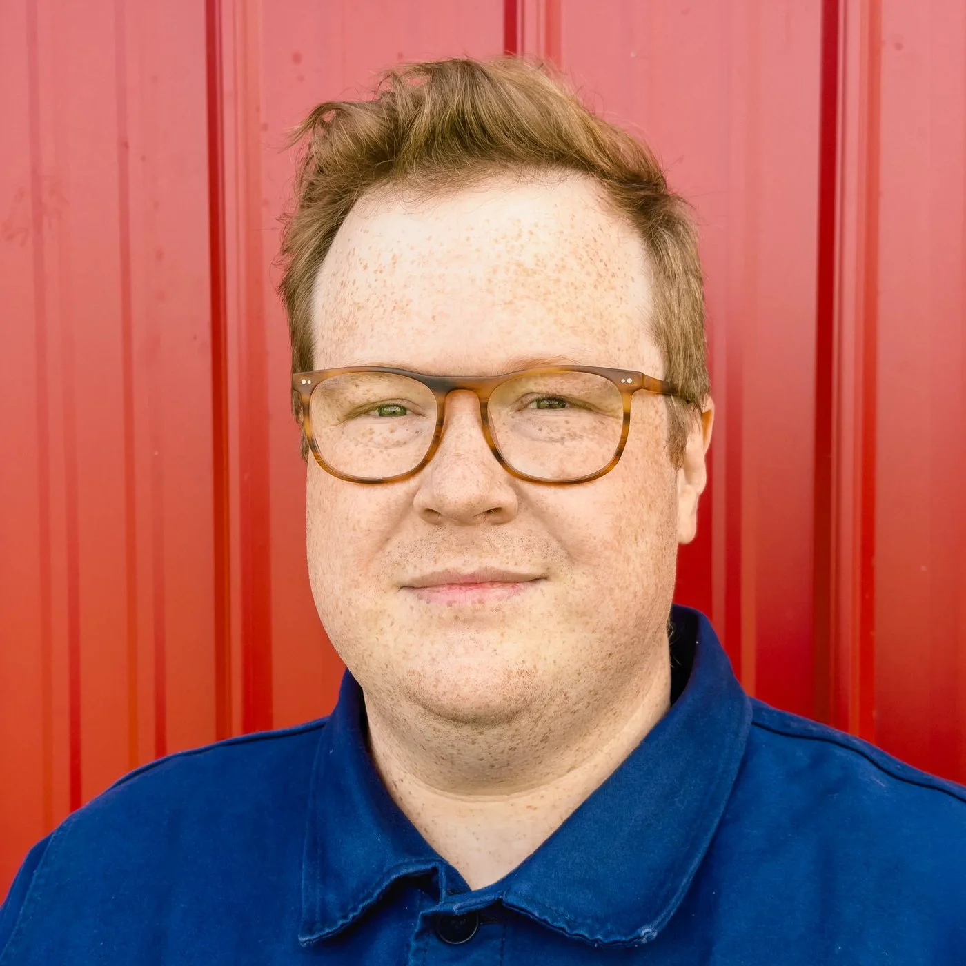 Sam Stringer-Hye in glasses and a navy work jacket, standing in front of a red corrugated wall